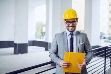 Smiling satisfied caucasian in gray suit with helmet on head and folder in hands posing outside.