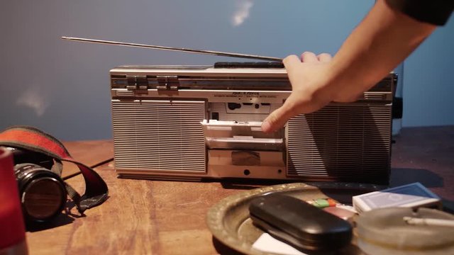 Cassette deck on radio opened and closed by hand, diverse objects beside it on table