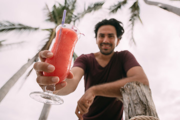 A handsome guy holds a fruit smoothie in his hands and smiles pretty on the background of palm trees.