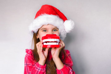 happy funny baby girl in red Christmas hat with toy model teeth on grey background.