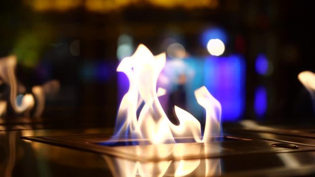 A slow motion close up of an open flame outside a restaurant to appeal to the higher class and the wealthy. Peoples silhouette / figures walk through the background to resemble a busy atmosphere.
