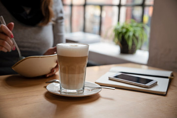 Latte coffee in a glass with smartphone on a wooden table in loft interior of cafe with window. Woman eats meal during lunch time. 
