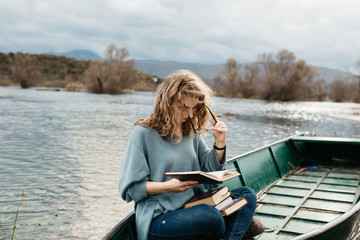 Fototapeta premium Portrait of young beautiful woman sitting on a boat and reading a book. She is bookworm and she choose between few books. 