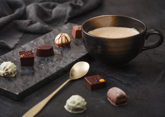 Luxury Chocolate candies selection with cup of cappuccino coffee and dessert spoon on black marble board and dark table background.