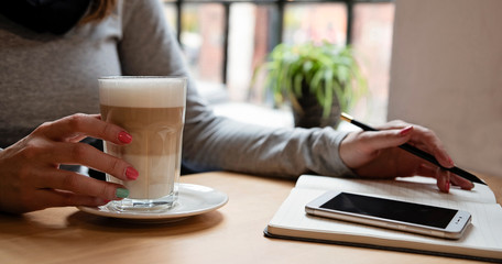 young woman working on her phone with notebook, pencil and coffee on a wooden table at home with window. Panoramic banner