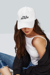 Cropped upward shot of a dark-haired girl, wearing white baseball cap with print with lettering "life is fantastic", white tank top and black jacket. The cap peak is closing upper part of a face. 