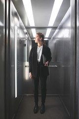 Vertical photo of young calm businesswoman holding laptop in her hand and looking at floor number in lift. Female office worker keeping laptop in her hand looking up in elevator. Full-length photo.