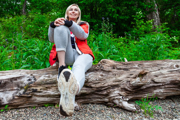 Beautiful blonde girl in a autumn jacket with a red backpack sits on a fallen tree in a forest or...