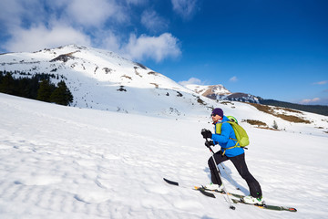 Fototapeta na wymiar Male tourist in colorful clothing and sunglasses with backpack trekking on skis in deep snow on background of bright blue sky and beautiful mountain. Winter vacations, ski touring concept.