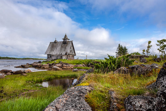 Russia, Karelia, White Sea, near Kem: Old rund down wooden Saint Nicholas chapel in rural remote natural place environment with calm lake water and dark blue cloudy sky - concept lost place outdoor