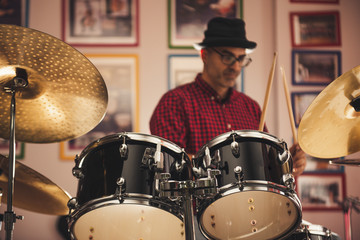 Caucasian young man with glasses and beret wearing a red and black plaid shirt, playing the drums with a soft side light
