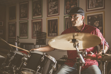 Caucasian young man with glasses and beret wearing a red and black plaid shirt, playing the drums with a soft side light