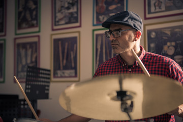 Caucasian young man with glasses and beret wearing a red and black plaid shirt, playing the drums with a soft side light