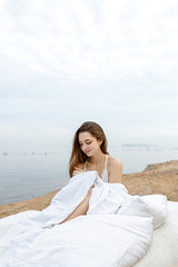 Fototapeta na wymiar Young slender woman sits on a white bed on a background of the sea and smiles