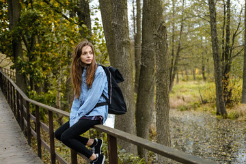 Woman tourist with a backpack sits on a bridge in the autumn forest.