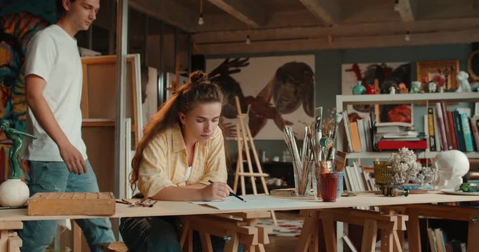 Caucasian young beautiful female painter workingin the art studio and drawing a sketch with a pencil when a man, her boyfriend, coming and telling his opinion on the picture.
