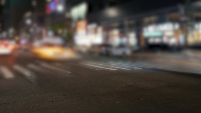 New York City traffic timelapse shot on a busy Broadway intersection at night, focus on asphalt, background blurred, long exposure. Shows yellow nyc taxis and pedestrians taking turns to pass.
