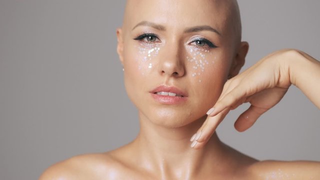 Cheerful young hairless woman with fashion eye makeup looking to the camera isolated over gray wall