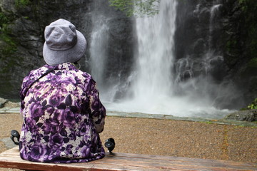 An old lady sitting on a bench and looking at waterfall