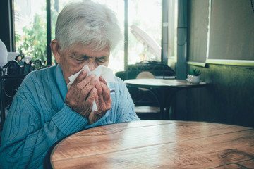 Old person coughing covering mouth with a tissue on a house interior. She has flu, allergy symptoms, acute bronchitis, pulmonary infections or pneumonia. Concept:respiratory infections and healthcare.