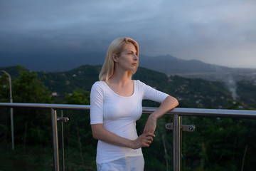 Blonde caucasian woman in standing near the edge of observation deck viewpoint or balcony with metallic handrails and transparent glass walls looking towards mountains in evening view from back