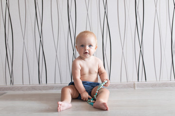 Little baby chewing her special chain toy for chewing made from silicone in blue jeans sitting on a white floor in a bright room inside apartments naked torso portrait looking in camera