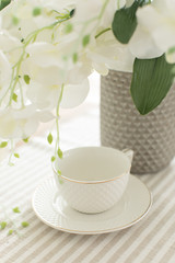 A white empty cup with plate on white tablecloth on white bright background with white flowers of orchid and green leaves like wedding morning