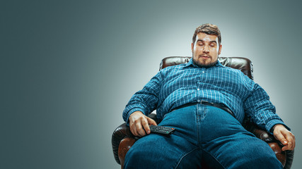Portrait of fat caucasian man wearing jeanse and whirt sitting in a brown armchair isolated on...
