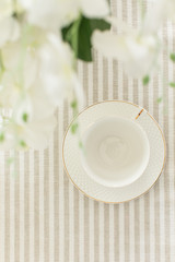 A white empty cup with plate on white tablecloth on white bright background with white flowers of orchid and green leaves like wedding morning