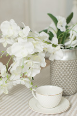 Obraz na płótnie Canvas A white empty cup with plate on white tablecloth on white bright background with white flowers of orchid and green leaves like wedding morning