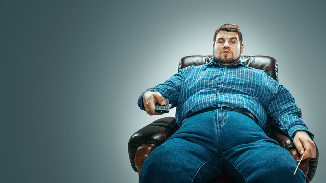 Portrait of fat caucasian man wearing jeanse and whirt sitting in a brown armchair isolated on gradient grey background. Emotional watching TV and changing channels, laughting. Overweight, carefree.