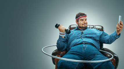 Look I'm getting sportive. Portrait of fat caucasian man sitting in a brown armchair on grey...