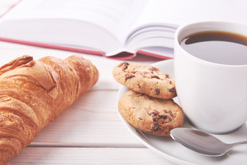 Still life with red book, cup of coffee or tea and two cookies. Croissant on white table top.