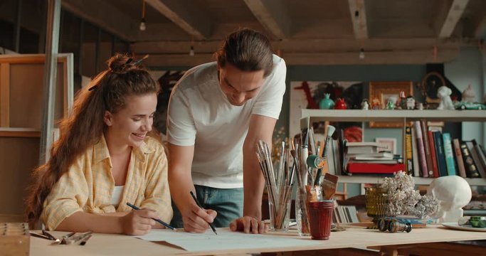 Young happy Caucasian good looking woman sitting at the desk and drawing with a pencil a sketch when her teacher, male artist, helping her, showing how to draw and teaching.
