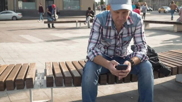 Man gray plaid shirt plaid shirt jeans is sitting on bench on street in the city In the hands of mobile phone He sends an SMS message Summer sunny day Passers by are coming Passing cars medium shot