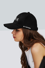 Cropped side photo of a girl, wearing black baseball cap with Chinese characters print and with lettering "be your muse" on the left side of cap, white tank top, black jacket and jeans with scuffs. 