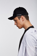 Cropped side photo of a dark-haired man, wearing black baseball cap with Chinese characters print and lettering "be your muse" on the left side of a cap, black t-shirt and white shirt. 