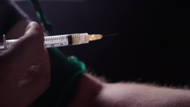 A man shooting heroin with a needle into his arm in a dark room