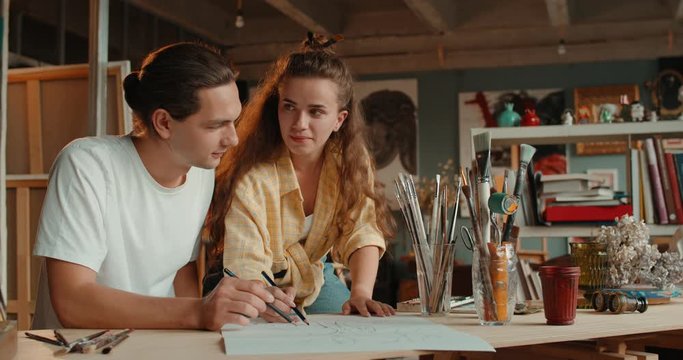 Happy young Caucasian couple of artists drawing together a sketch with pencils and smiling to each other in the cozy workshop.