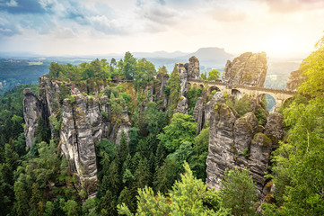 Beautiful panoramic view of famous Bastei Bridge with Elbe Sandstone mountains in Saxon Switzerland National Park with dark clouds in golden morning light at sunrise in spring, Saxony, Germany