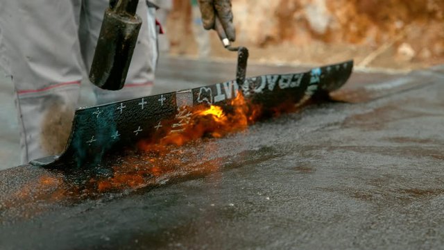 SLOW MOTION, CLOSE UP: Unrecognizable laborer lays black tar insulation over the building's foundation. Man runs a fire extinguisher along a sheet of bitumen coating being melted onto the ground.