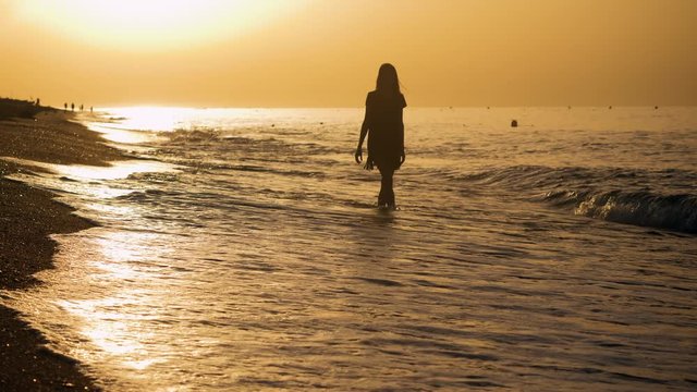 Coastline Costa del Maresme. Young beautiful woman walking barefoot across the sandy beach of Mediterranean sea at sunrise. Spain. 4K