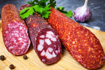 Three types of delicious smoked sausages on a wooden cutting board. Nearby are peppercorns, parsley and garlic. Dark marble background.