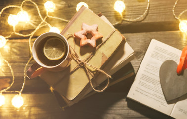Holidays mood photo. Christmas lights and hot tea mug. Book for cosy evening. Sweet gingerbread and wooden heart on tray. Perfect winter flat lay with candle. Hygge concept.
