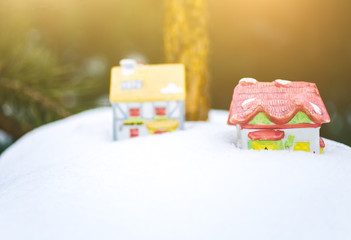 Original ceramic toy houses standing in snow on Christmas pine tree branch. Natural photo of decorations in frozen forest. Happy holidays ornaments postcard.