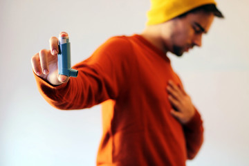 Man dressed in orange and wearing a yellow cap using a blue asthma inhaler to prevent an asthma attack. - obrazy, fototapety, plakaty