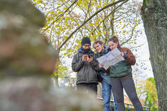 Some friends orienting themselves in a city, one with the smartphone and the others with a paper map, in the sky the yellow eyes of a tree are seen, in the foreground there are some unfocused stones