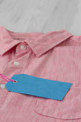 Blank blue tag label on a mens pink shirt with white buttons.  On a grey wood background