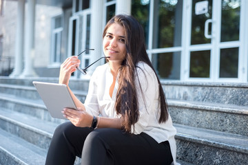 Fototapeta premium portrait of a beautiful indian girl. Business woman smiles, holding glasses, a notebook, sits on the steps of an office building