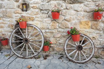 Fototapeta na wymiar Unusual street decor in Greece. Two old vintage wooden wheels standing near stony house wall decorated with many buckets with growing flowers. Horizontal color photography.